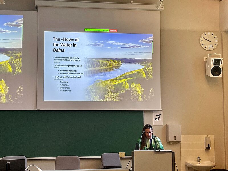 In the picture, a presentation slide with an image of the river and forest landscape on the right and the title "The How of the Water in Daina". Presented by lead researcher Anne Sauka, who is located on the lower right edge of the picture. 