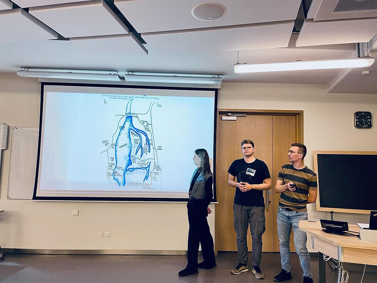 Attēla kreisajā malā atrodas prezentācijas ekrāns ar slaidu, kurā attēlota zīmēta karte ar ūdenstilpni. Labajā malā - trīs studenti, kuri veic prezentēšanu. Attēla kreisajā malā atrodas prezentācijas ekrāns ar slaidu, kurā attēlota zīmēta karte ar ūdenstilpni. Labajā malā - trīs studenti, kuri veic prezentēšanu.