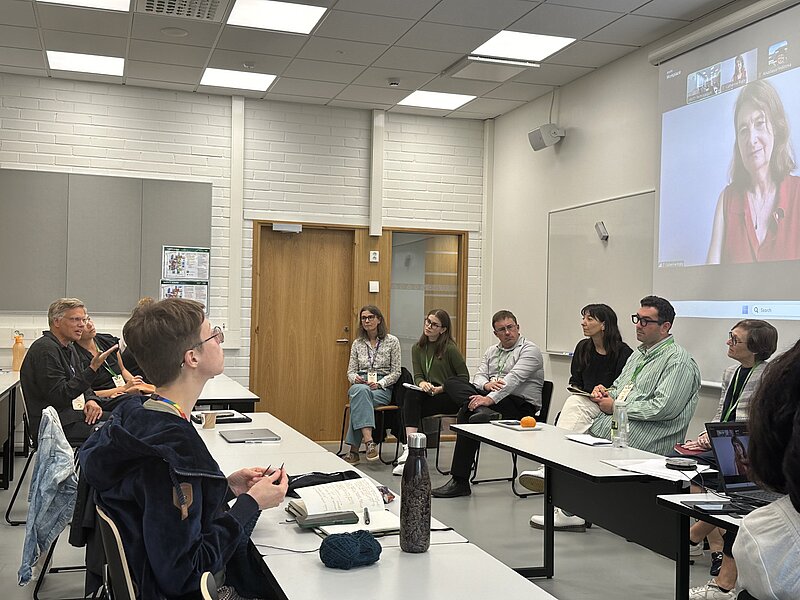 In the picture, a classroom where more than 10 conference participants sit at several white tables. On the upper right edge of the picture, a presentation screen showing a woman in a red dress and in smaller windows other conference participants connected remotely. 