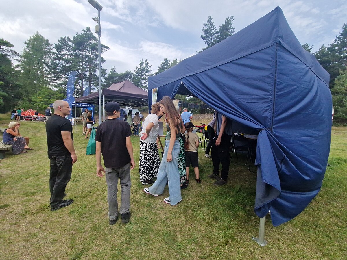 Pie tumši zilas āra pasākuma nojumes pulcējušies vairāki cilvēki, daļa sarunājas pie tās ieejas. Nojume atrodas zālienā līdzās citām pasākuma teltīm, apkārt redzami arī citi apmeklētāji. Pie tumši zilas āra pasākuma nojumes pulcējušies vairāki cilvēki, daļa sarunājas pie tās ieejas. Nojume atrodas zālienā līdzās citām pasākuma teltīm, apkārt redzami arī citi apmeklētāji.