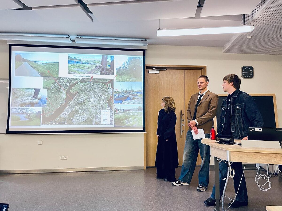 Attēla kreisajā pusē redzams prezentācijas ekrāns ar satelītattēliem un citiem foto. Labajā pusē atrodas trīs studenti, kuri prezentē. Attēla kreisajā pusē redzams prezentācijas ekrāns ar satelītattēliem un citiem foto. Labajā pusē atrodas trīs studenti, kuri prezentē.