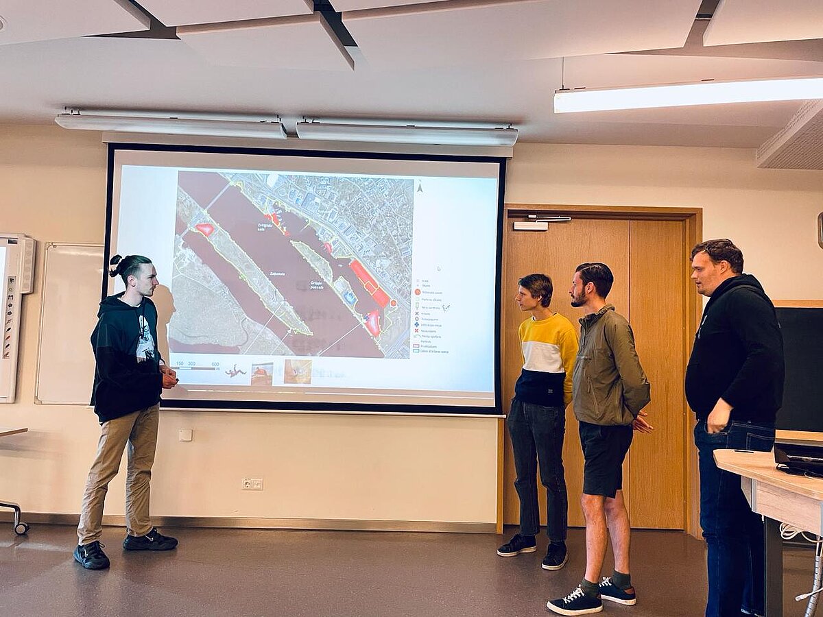 Attēla kreisajā malā redzams prezentācijas ekrāns ar slaidu, kurā redzama satelītattēls. Viens no studentiem atrodas ekrāns kreisajā malā, trīs pārējie - labajā malā. Attēla kreisajā malā redzams prezentācijas ekrāns ar slaidu, kurā redzama satelītattēls. Viens no studentiem atrodas ekrāns kreisajā malā, trīs pārējie - labajā malā.