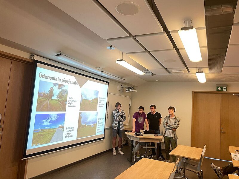 The picture shows the classroom where the presentation is taking place. You can see a slide titled "Waterfront Accessibility" presented by four students.