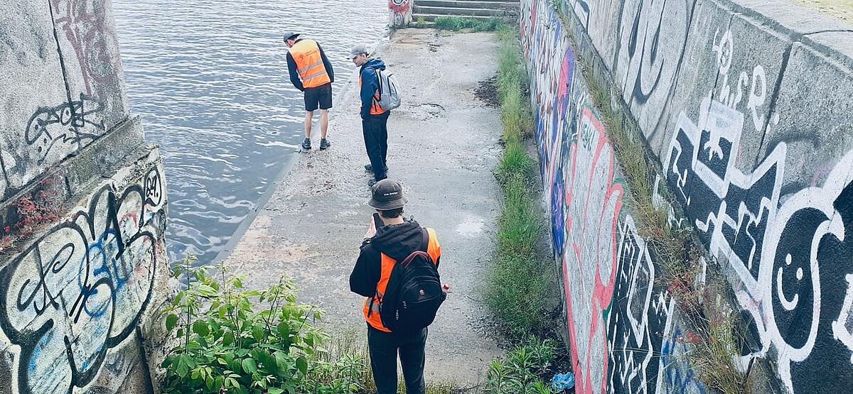 Attēlā redzami trīs studenti, kuri atrodas pie betonētas ūdenstilpnes malas. Betona sienas ir apzīmētas ar dažādu krāsu grafiti zīmējumiem. Attēlā redzami trīs studenti, kuri atrodas pie betonētas ūdenstilpnes malas. Betona sienas ir apzīmētas ar dažādu krāsu grafiti zīmējumiem.