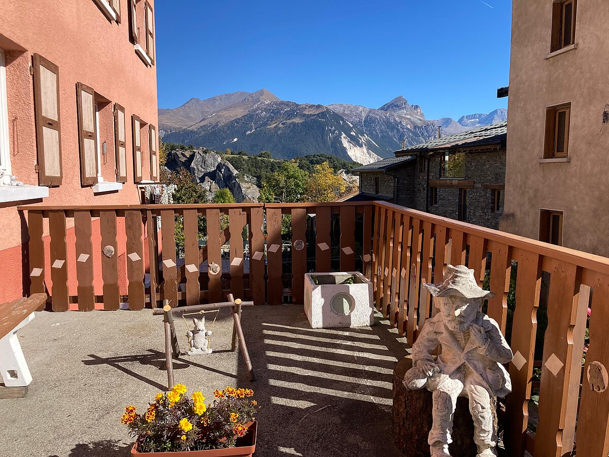 Attēlā redzams skats uz terasi, uz kuras izvietas dārza figūras. Fonā redzamas māju sienas un kalni. Attēlā redzams skats uz terasi, uz kuras izvietas dārza figūras. Fonā redzamas māju sienas un kalni.