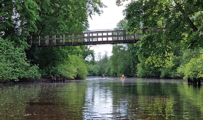 The picture shows a river surrounded by trees and bushes on both sides. Two orange canoe-type boats with boatmen can be seen further down the river. A gray wooden bridge stretches across the river.