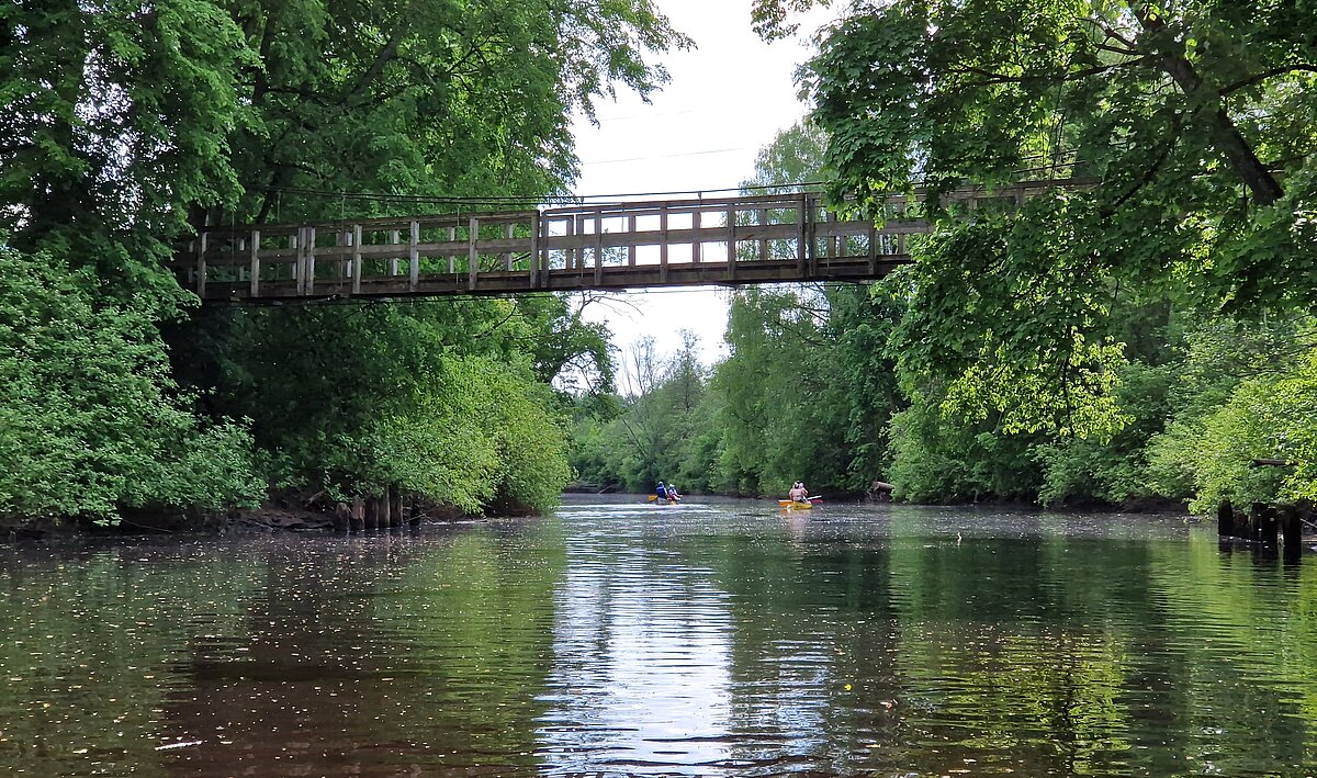 A river with a canoe and two canoeists. The river is surrounded on both sides by trees and bushes. Two orange canoes with boats are seen in the river below. A grey wooden bridge spans the river. A river with a canoe and two canoeists. The river is surrounded on both sides by trees and bushes. Two orange canoes with boats are seen in the river below. A grey wooden bridge spans the river.