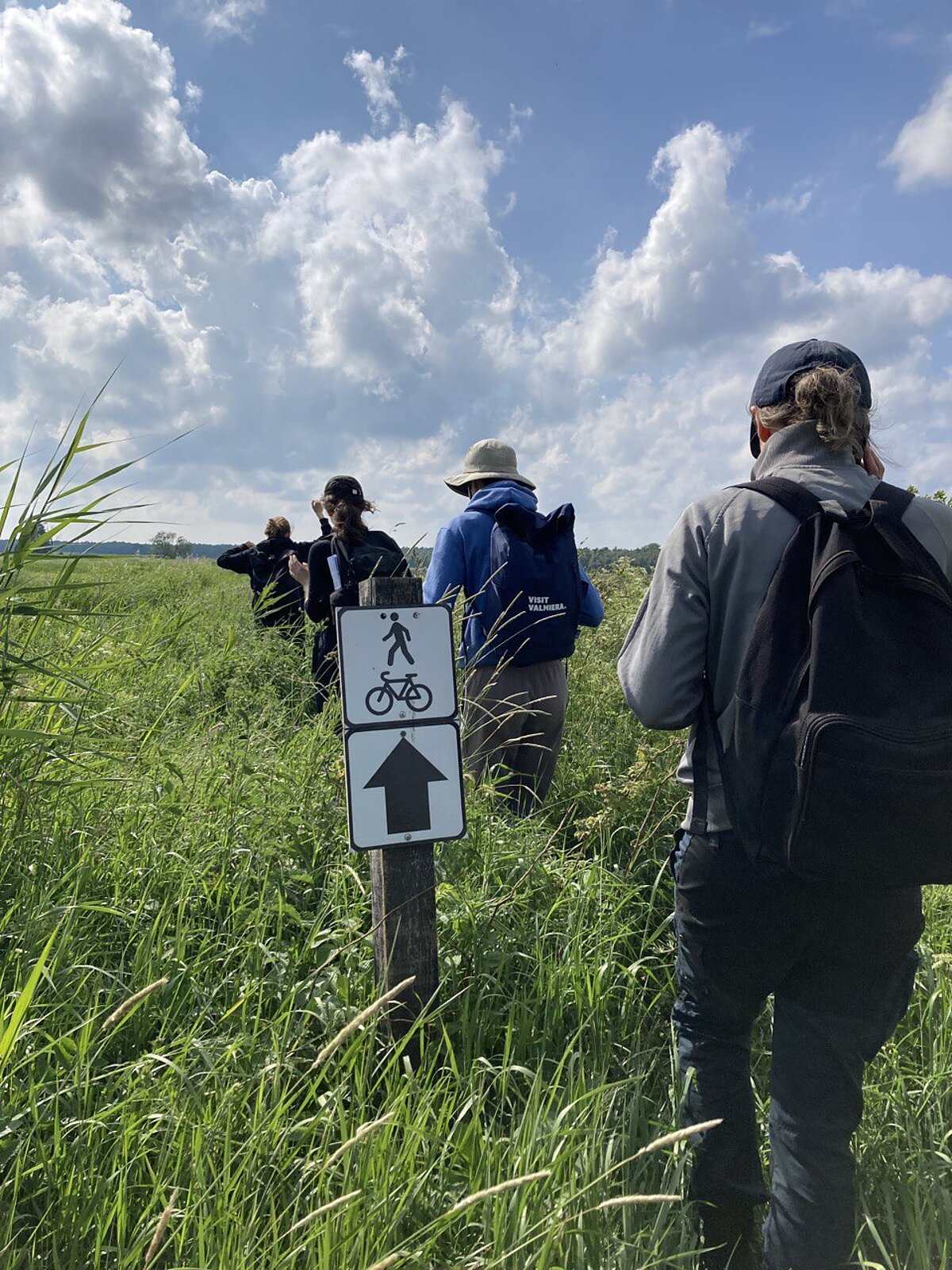 The group walks along a narrow, overgrown trail that is very closely surrounded by evergreens. A sign with an arrow, a pedestrian and a bicycle can be seen by the trail. The group walks along a narrow, overgrown trail that is very closely surrounded by evergreens. A sign with an arrow, a pedestrian and a bicycle can be seen by the trail.