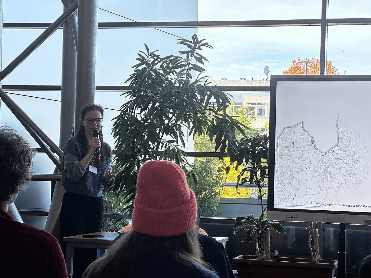 Attēla kreisajā daļā redzama projekta pētniece Kristīne Krumberga. Labajā pusē - daļa no prezentācijas slaida ar Latvijas kontūru un upju tīklu. Fonā telpaugs, stikla siena un blakus esoša ēka. Priekšplānā klausītāji. Attēla kreisajā daļā redzama projekta pētniece Kristīne Krumberga. Labajā pusē - daļa no prezentācijas slaida ar Latvijas kontūru un upju tīklu. Fonā telpaugs, stikla siena un blakus esoša ēka. Priekšplānā klausītāji.