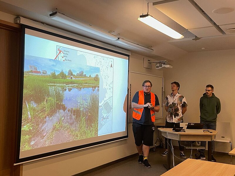 Attēlā mācību telpa, kurā notiek prezentācija. Redzams slaids ar ūdenstilpes attēli un karti fonā. Prezentē trīs studenti.