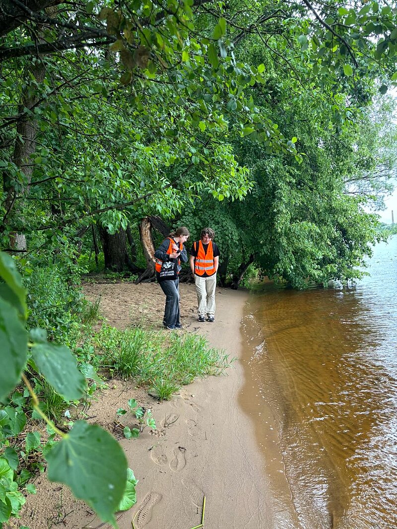 The picture shows the water's edge. Two students in orange vests are standing next to it. Trees, bushes and other plants on the edges of the picture.