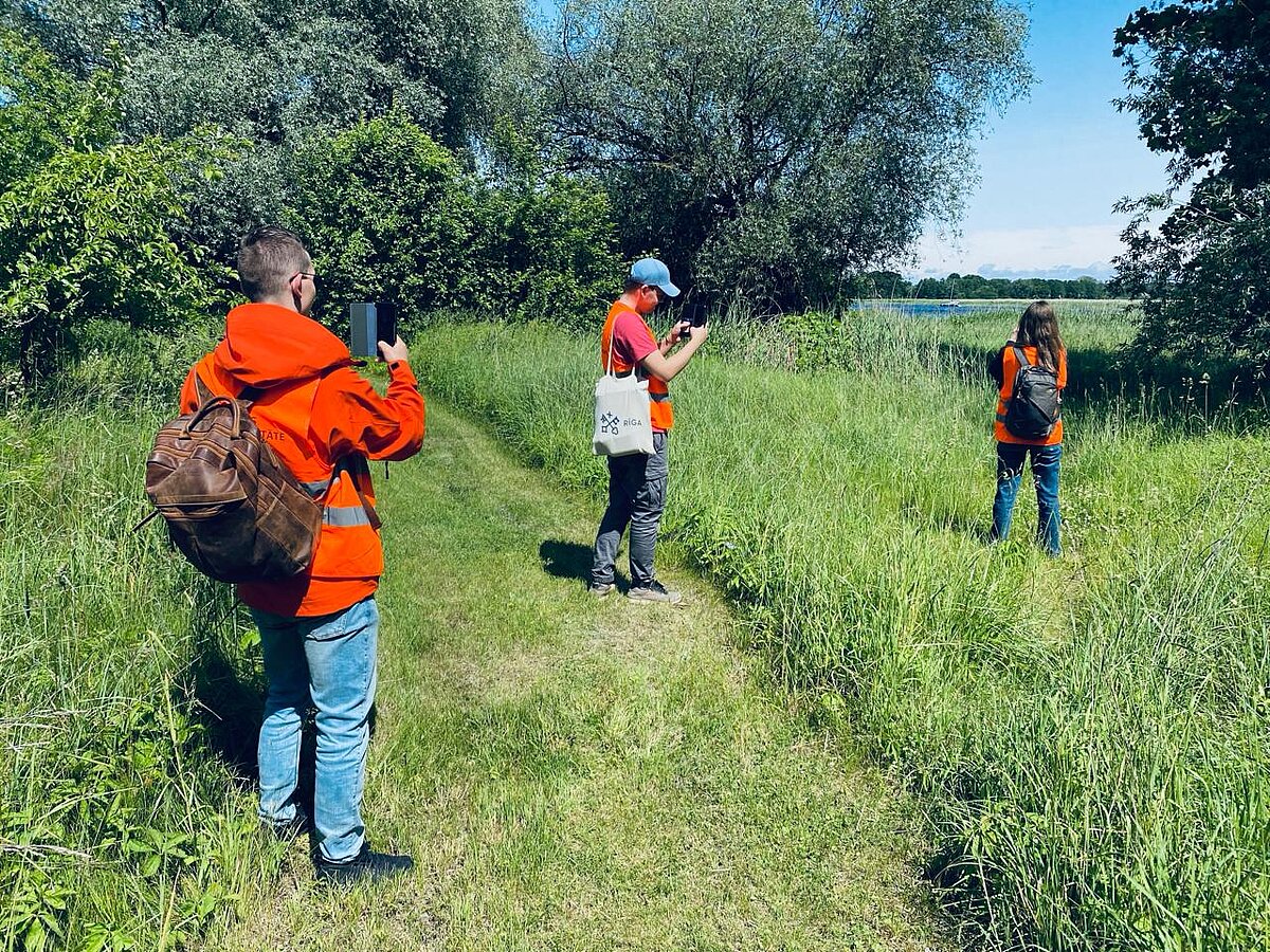 Attēlā redzami trīs studenti, kuri viens otru fotografē, atrodoties apaugušā ūdenstipnes krastā. Attēlā redzami trīs studenti, kuri viens otru fotografē, atrodoties apaugušā ūdenstipnes krastā.