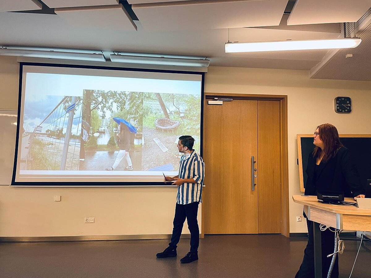 Kreisajā pusē atrodas prezentācijas ekrāns ar slaidu, kurā redzami trīs attēli. Viens no studentiem atrodas blakus ekrānam, otrs students - pie prezentēšanas pults. Kreisajā pusē atrodas prezentācijas ekrāns ar slaidu, kurā redzami trīs attēli. Viens no studentiem atrodas blakus ekrānam, otrs students - pie prezentēšanas pults.