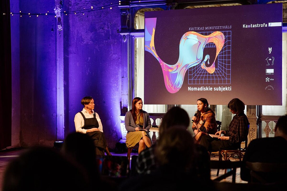 Attēla centrālajā daļā redzamas diskusijas dalībnieces, no kreisās - Dace Bula, Ieva Astahovska, Anne Sauka, Anita Zariņa. Attēla augšējā labajā daļā redzams prezentācijas ekrāns ar uzrakstiem "Kritikas minifestivāls. Nomadiskie subjekti. Kantastrofa". Attēlā priekšplānā redzami daļa no diskusijas klausītājiem. Attēla centrālajā daļā redzamas diskusijas dalībnieces, no kreisās - Dace Bula, Ieva Astahovska, Anne Sauka, Anita Zariņa. Attēla augšējā labajā daļā redzams prezentācijas ekrāns ar uzrakstiem "Kritikas minifestivāls. Nomadiskie subjekti. Kantastrofa". Attēlā priekšplānā redzami daļa no diskusijas klausītājiem.