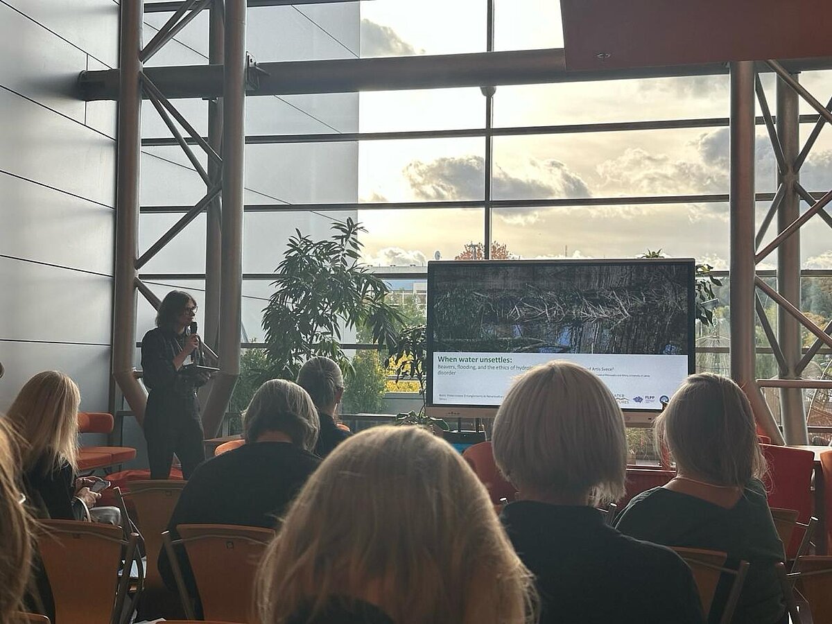 Attēla kreisajā pusē redzama projekta pētniece Anita Zariņa. Labajā un centrālajā daļā - prezentācijas ekrāns ar virsrakstu "When water unsettles". Fonā stikla siena, telpaugi un citas blakusēkas. Priekšplānā - prezentācijas klausītāji. Attēla kreisajā pusē redzama projekta pētniece Anita Zariņa. Labajā un centrālajā daļā - prezentācijas ekrāns ar virsrakstu "When water unsettles". Fonā stikla siena, telpaugi un citas blakusēkas. Priekšplānā - prezentācijas klausītāji.