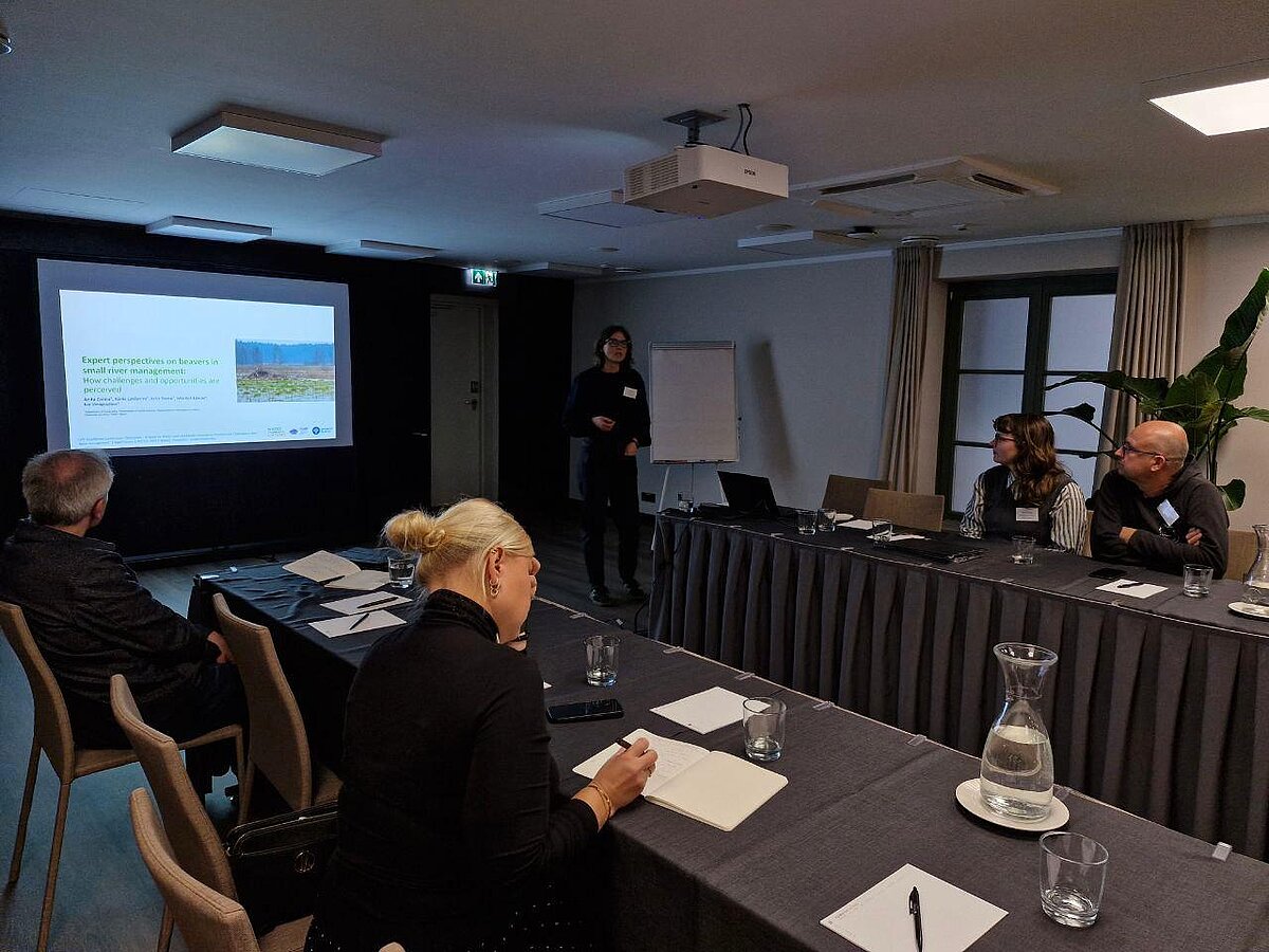 Attēlā redzama referāta prezentēšana. Kreisajā pusē redzams prezentācijas slaids ar tekstu. Priekšplānā un labajā malā galds, ap kuru sēž cilvēki. Attēla centrālajā, tālakajā daļā redzama projekta pētniece Anita Zariņa. Attēlā redzama referāta prezentēšana. Kreisajā pusē redzams prezentācijas slaids ar tekstu. Priekšplānā un labajā malā galds, ap kuru sēž cilvēki. Attēla centrālajā, tālakajā daļā redzama projekta pētniece Anita Zariņa.