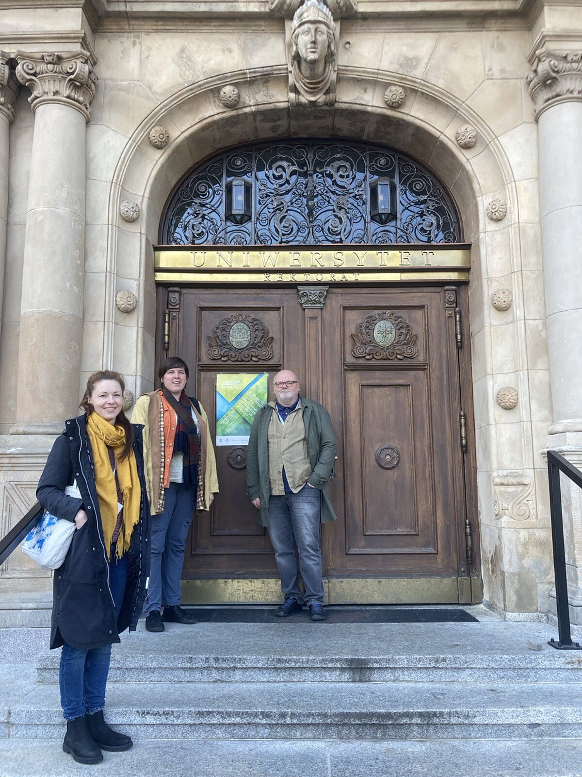 In the background of the picture, a brown wooden door in the building, at which three researchers of the project are standing. From the left - Kristīne Krumberga, Karīna Ješkina and Artis Svece. The inscription "Universytet Rektotat" on a gilded plate above the door. In the background of the picture, a brown wooden door in the building, at which three researchers of the project are standing. From the left - Kristīne Krumberga, Karīna Ješkina and Artis Svece. The inscription "Universytet Rektotat" on a gilded plate above the door.