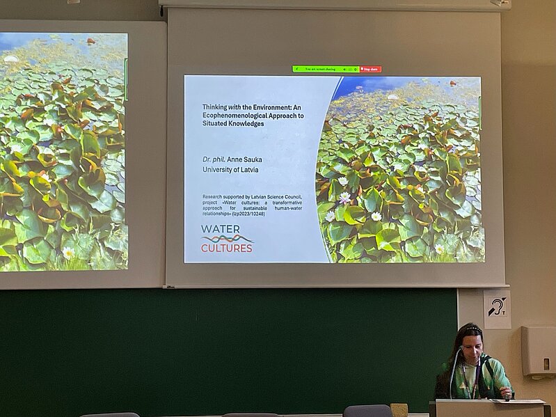 In the picture, a presentation slide with an image of water lilies on the right and the title "Thinking with the Environment: An ecophenomenological Approach to Situated Knowledges". Presented by leading researcher Anne Sauka, who is located on the lower right edge of the picture. 