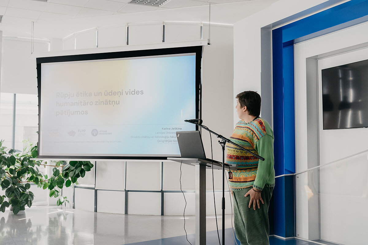 On the right side of the image is the author of the presentation, Karīna Ješkina. In the center and on the left is the presentation screen with the title slide and the inscription "Ethics of Care and Water in Environmental Humanities." On the right side of the image is the author of the presentation, Karīna Ješkina. In the center and on the left is the presentation screen with the title slide and the inscription "Ethics of Care and Water in Environmental Humanities."