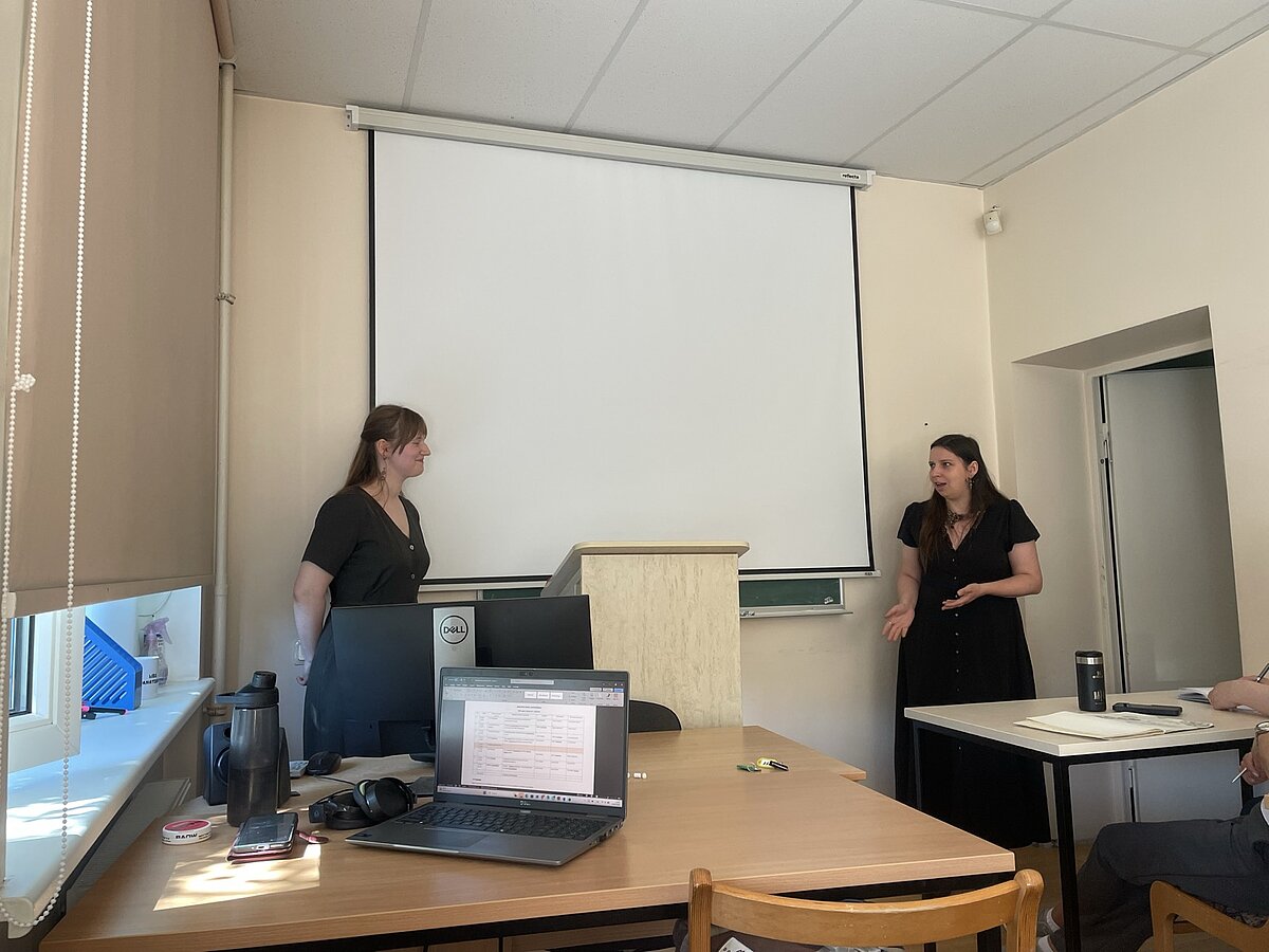 Paula Lūcija Lejiņa uzstājas pie lektora pults auditorijā. Fonā redzams projekcijas ekrāns, bet priekšplānā klausītāji un galdi ar datoriem, radot akadēmiskas nodarbības atmosfēru. Paula Lūcija Lejiņa uzstājas pie lektora pults auditorijā. Fonā redzams projekcijas ekrāns, bet priekšplānā klausītāji un galdi ar datoriem, radot akadēmiskas nodarbības atmosfēru.