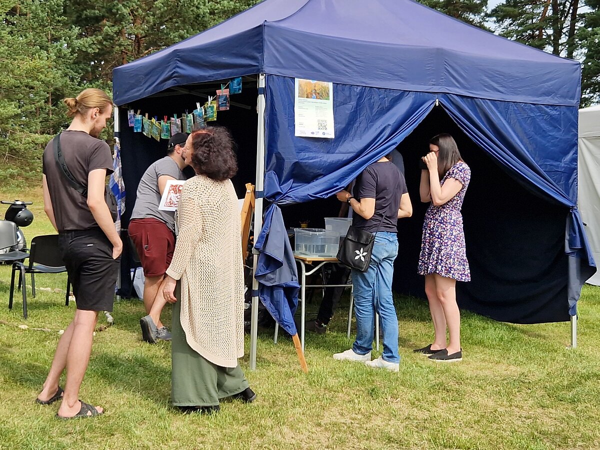 A group of visitors and project participants gather around the dark blue festival tent. Transparent containers used for an activity are placed on a table near the entrance, with an illustration displayed on an easel nearby. A group of visitors and project participants gather around the dark blue festival tent. Transparent containers used for an activity are placed on a table near the entrance, with an illustration displayed on an easel nearby.