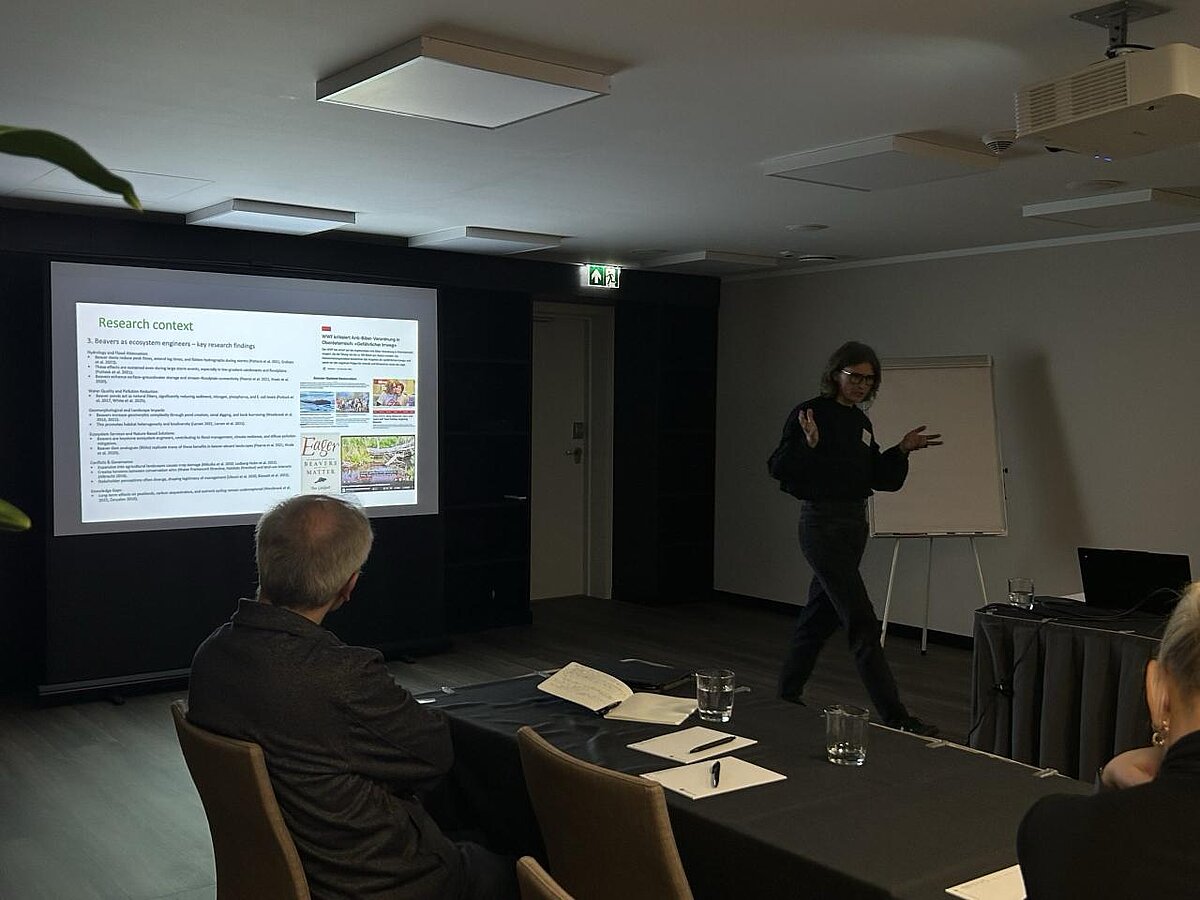 The picture shows a presentation of a report. On the left side, you can see a presentation slide with text. In the foreground, there is a table with people sitting around it. On the right side, there is project researcher Anita Zariņa. The picture shows a presentation of a report. On the left side, you can see a presentation slide with text. In the foreground, there is a table with people sitting around it. On the right side, there is project researcher Anita Zariņa.