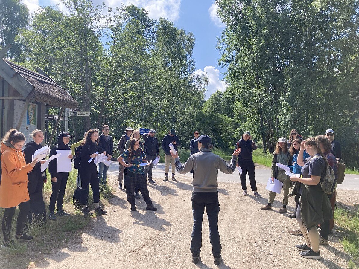 Students stand in a semicircle around the professor, who presents the tasks. Trees and camping log cabins can be seen in the background. Students stand in a semicircle around the professor, who presents the tasks. Trees and camping log cabins can be seen in the background.