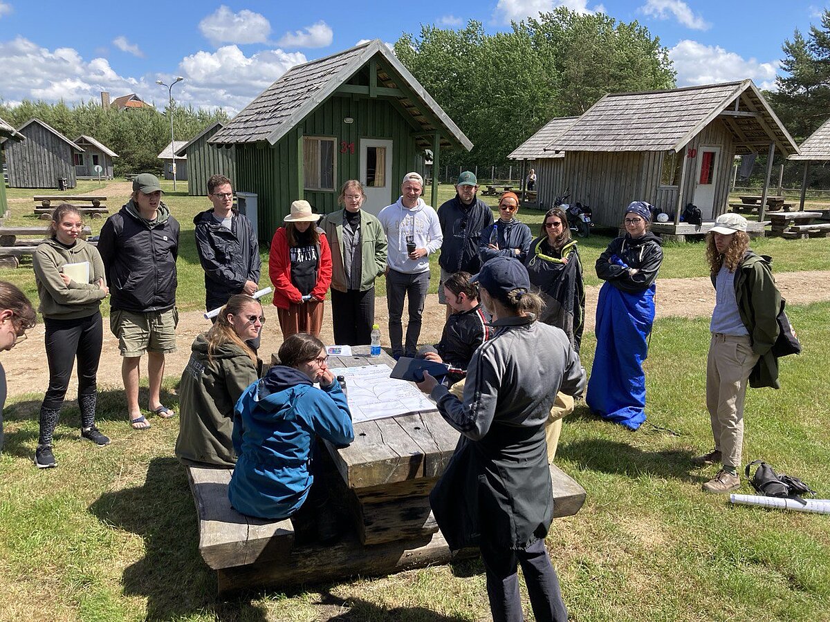 Group work presentation. Students sit at a wooden table and present the scheme and drawings drawn on an A3 sheet. In the background of the picture are listeners and wooden houses. Group work presentation. Students sit at a wooden table and present the scheme and drawings drawn on an A3 sheet. In the background of the picture are listeners and wooden houses.