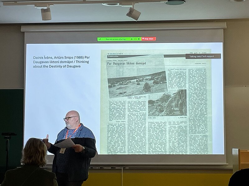 In the picture, a presentation slide with the text "Dainis Īvāns, Artūrs Snips (1986) Thinking about the fate of the Daugava" on the right and a newspaper article on the left, presented by the leading researcher Artis Svece, located on the left side.