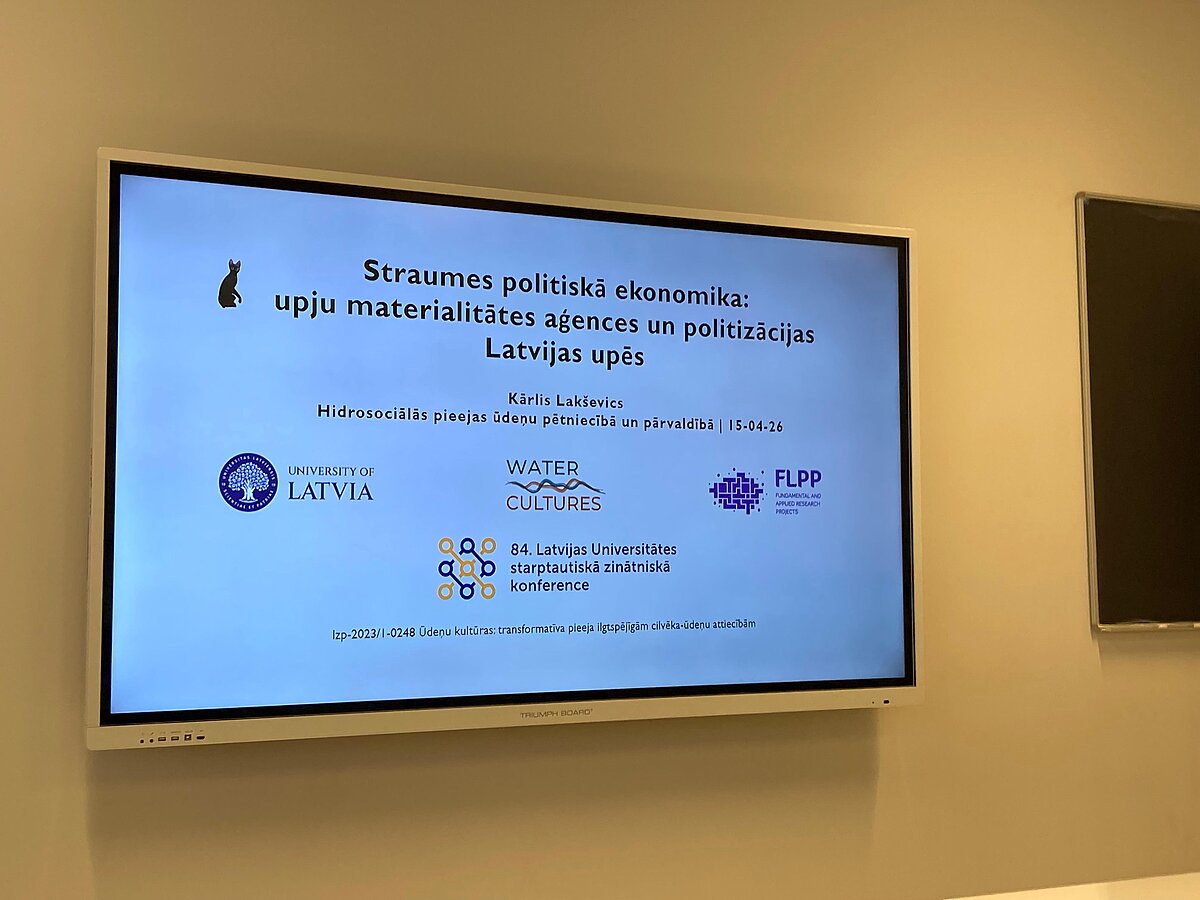 Attēlā redzams prezentācijas ekrāns ar titulslaidu. Prezentācijas nosaukums ir "Straumes politiskā ekonomika: upju materialitātes aģences un politizācijas Latvijas upēs". Attēlā redzams prezentācijas ekrāns ar titulslaidu. Prezentācijas nosaukums ir "Straumes politiskā ekonomika: upju materialitātes aģences un politizācijas Latvijas upēs".