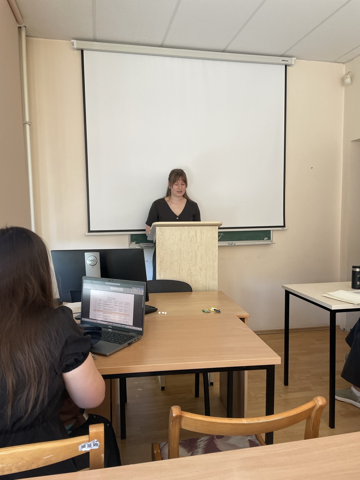 Paula Lūcija Lejiņa speaks from the lectern in a classroom. A projection screen is visible behind her, while audience members, desks, and laptops in the foreground create the atmosphere of an academic session. Paula Lūcija Lejiņa speaks from the lectern in a classroom. A projection screen is visible behind her, while audience members, desks, and laptops in the foreground create the atmosphere of an academic session.